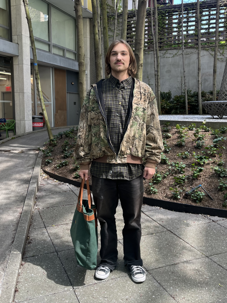 Student stands in Lang courtyard wearing a camouflage hoodie, plaid button-up shirt, black jeans, Converse sneakers, and a green tote bag.