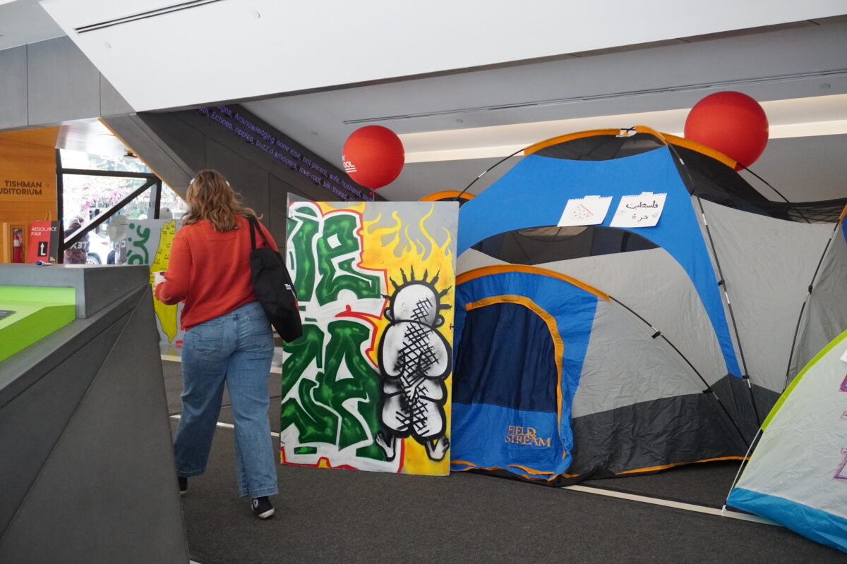 blue and grey tent on right side of photo next to multicolor sign with green lettering and a person in a red sweater and jeans is walking past on right side of the tent 