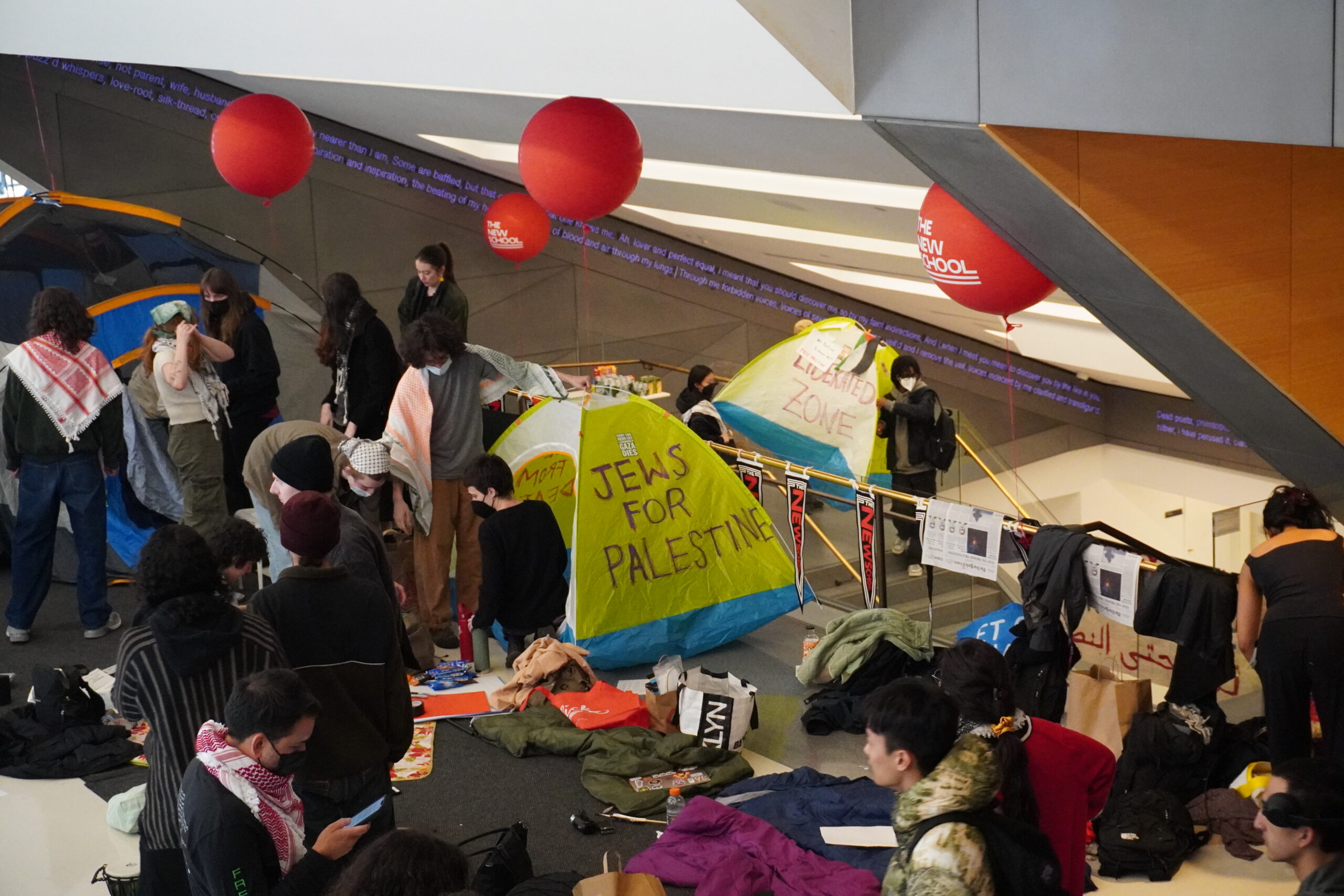 Multiple students walk amongst tents set up in the University Center lobby. In the background are red New School balloons and in the center of the image is a yellow tent that reads “Jews for Palestine.”