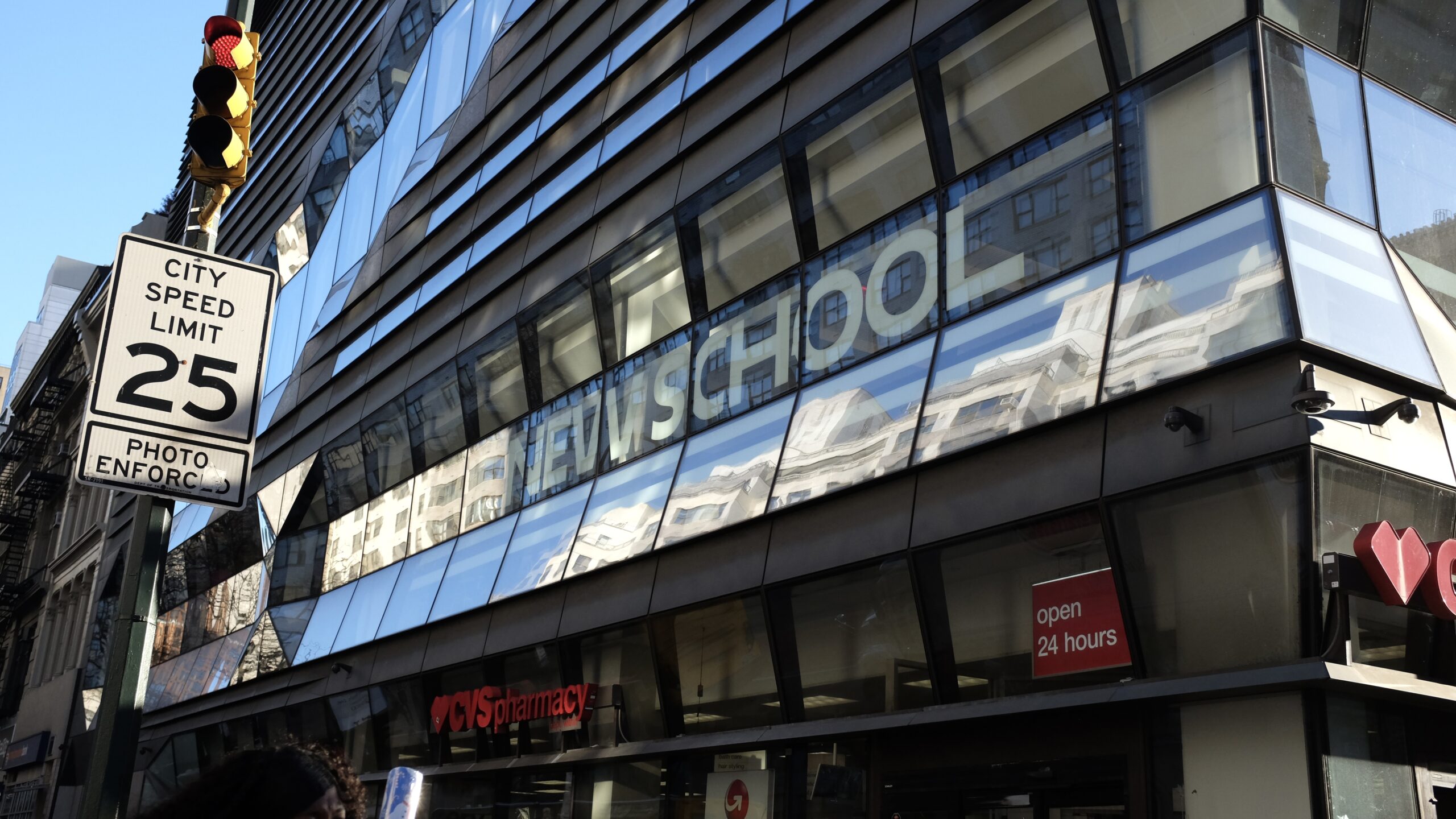 Image of The front of the New School’s University Center with school logo above CVS store, speed limit sign and stop light seen on the left side of image.