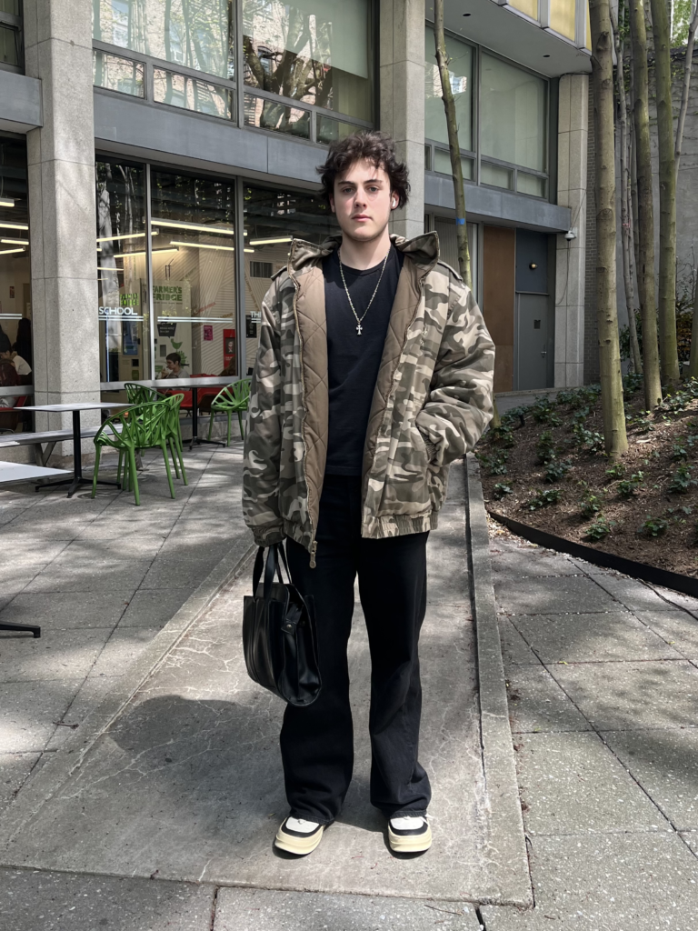 Student stands in Lang courtyard wearing a camouflage hoodie, a black T-shirt and jeans, black and white sneakers, a black tote bag, and a silver necklace.