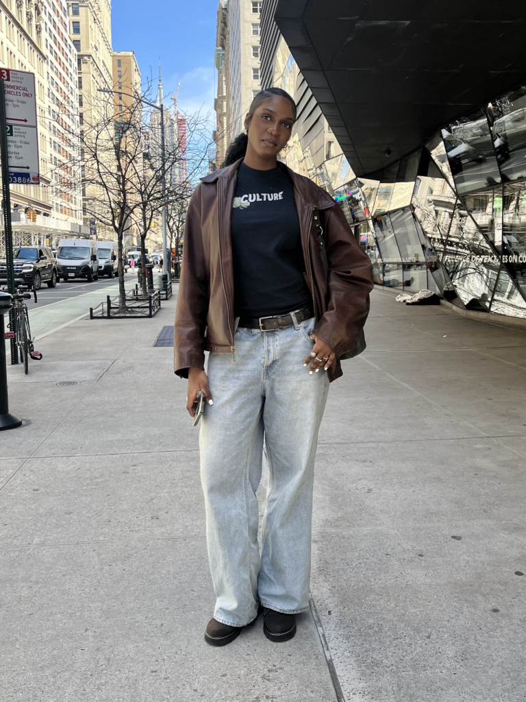 Student stands in front of The New School University Center wearing a black graphic t-shirt, brown leather jacket, light wash jeans, and brown boots.