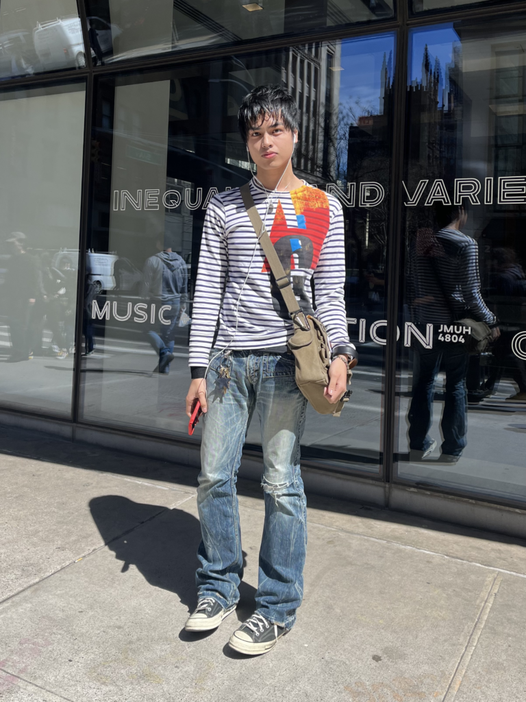 Student stands in front of The New School University Center wearing a striped graphic long sleeve, acid wash jeans, black Converse, and a brown messenger bag.
