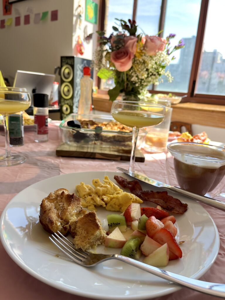 Plate of bread pudding, fruit salad, bacon, and eggs with a glass of mimosa.