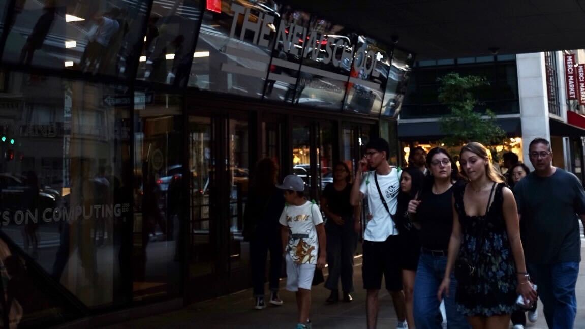 people walking on left side of photo in front of a dark building with three sets of doors