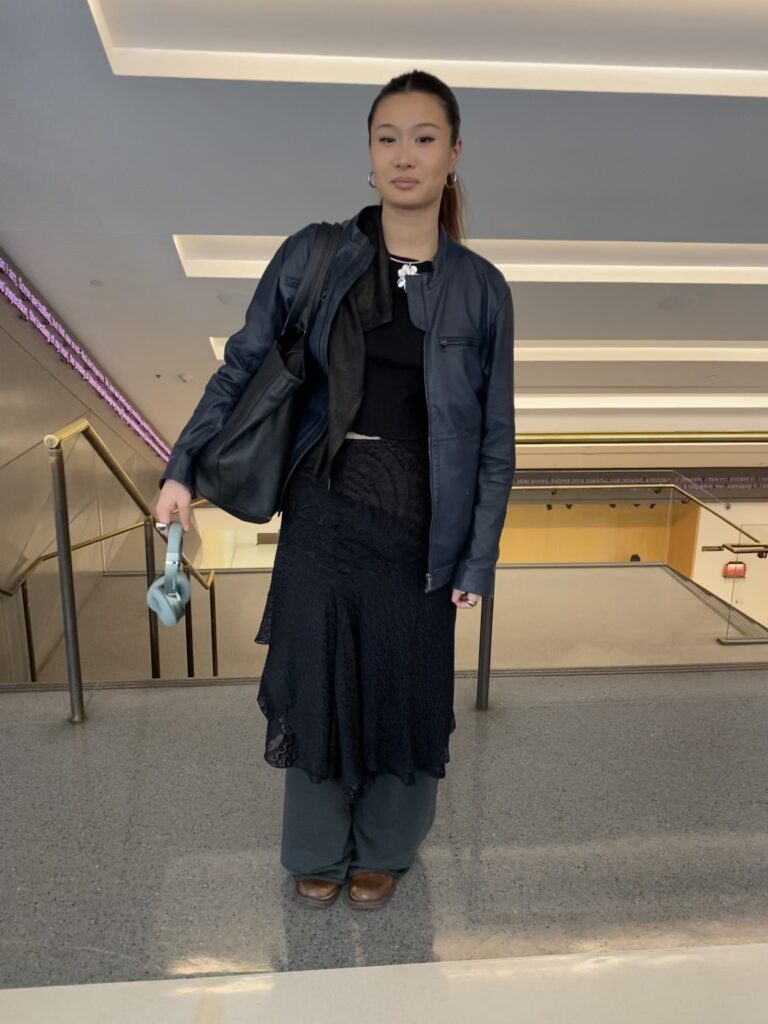 A student stands in the UC and wears a leather jacket over a black T-shirt, a textured black lace skirt over her sweatpants, and brown boots. She has a silver flower pendant on, a black bag on her shoulder, and holds headphones in her hand.
