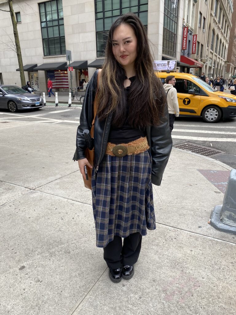 A student wears a blue tartan skirt over black pants, a chunky brown belt, a black leather jacket, brown leather bag, and patent leather shoes.