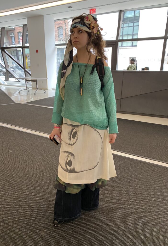 A student stands in the UC and wears a headscarf with flowers in it, two long necklaces with pendants, an off-the-shoulder teal sweater, and a skirt with a face on it layered over a green skirt and pinstripe pants.