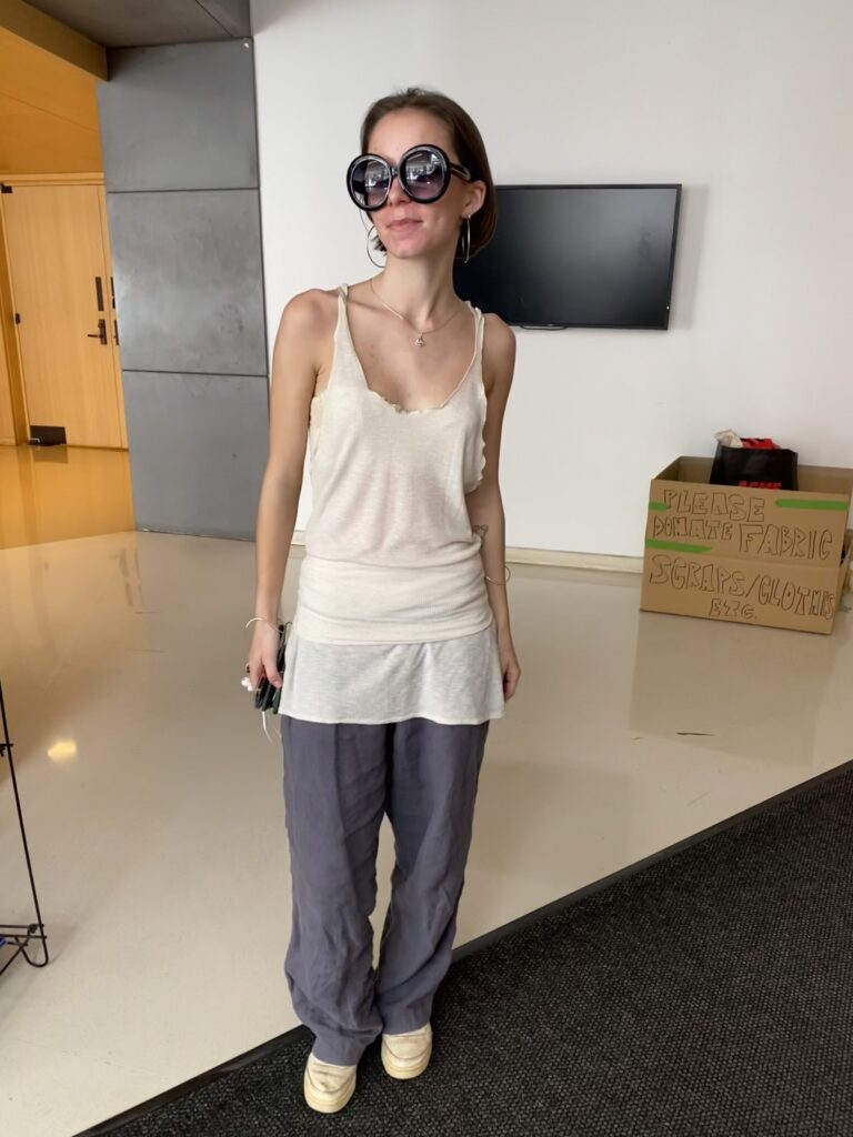 A student wears black circular sunglasses, a long, white, ribbed tank top folded into a skirt over gray pants, and white sneakers