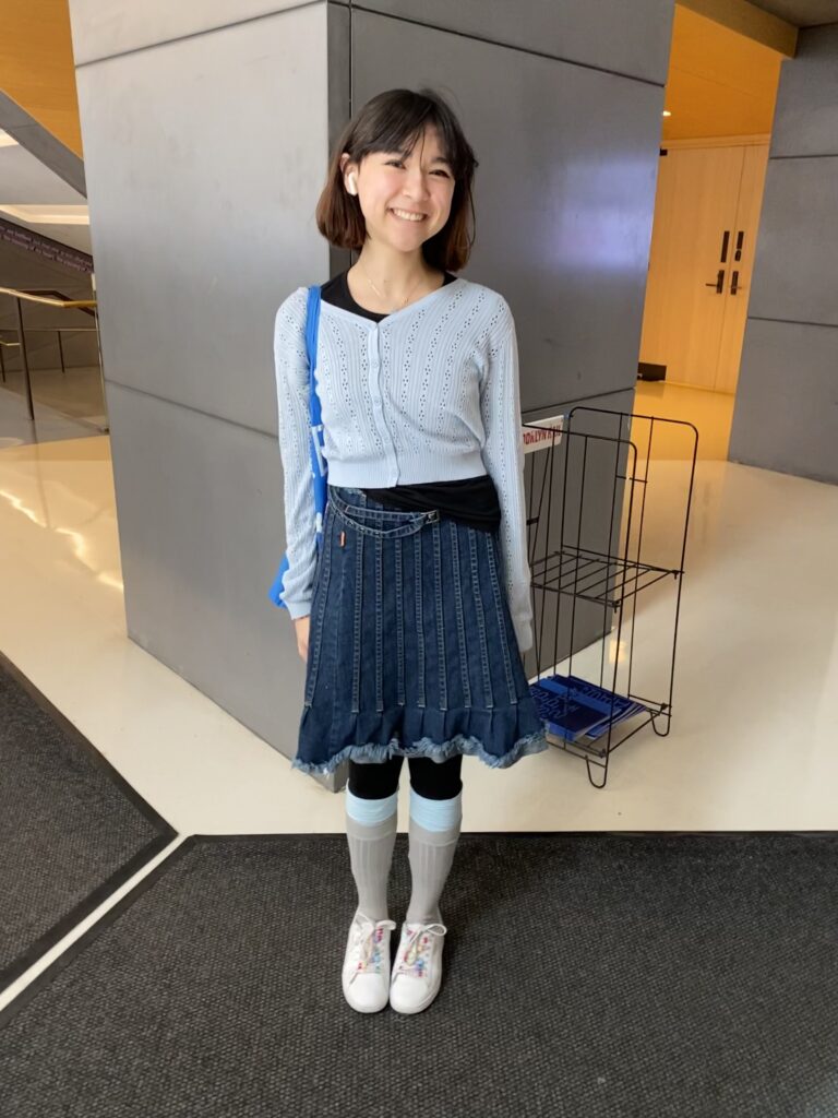 A student wears a light blue cardigan over a black shirt, a denim skirt with an uneven hemline, black leggings, light blue and gray under-the-knee socks, and white sneakers