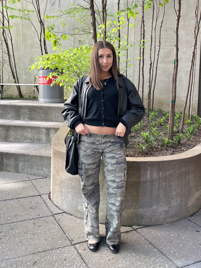 Student stands in Lang courtyard wearing camouflage cargo pants, a black cardigan, leather jacket, tote bag, and ballet flats.