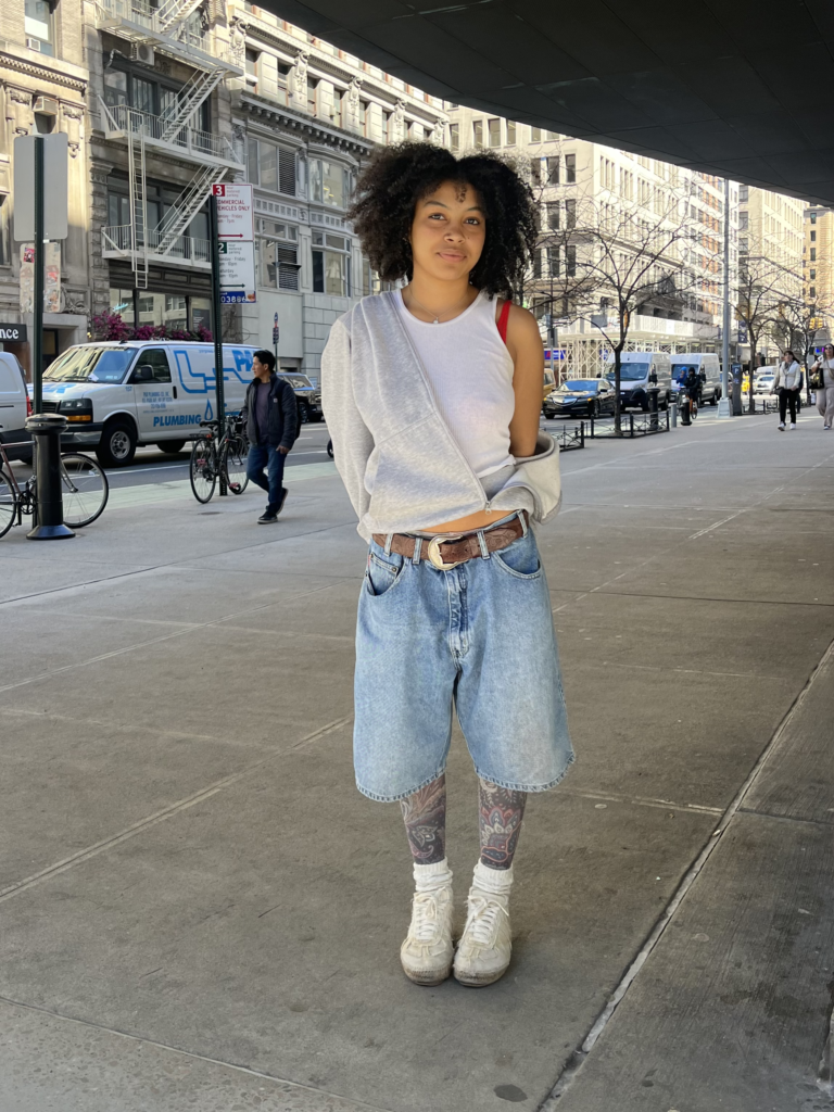 Student stands on Fifth Avenue wearing a white tank top, gray hoodie, brown leather belt, baggy jean shorts, graphic tights, and white sneakers.