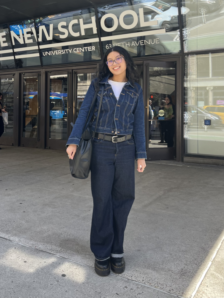 Student stands in front of The New School University Center wearing glasses, a white t-shirt, dark wash denim jacket, dark wash jeans, black platform boots, and a black Telfar tote bag.