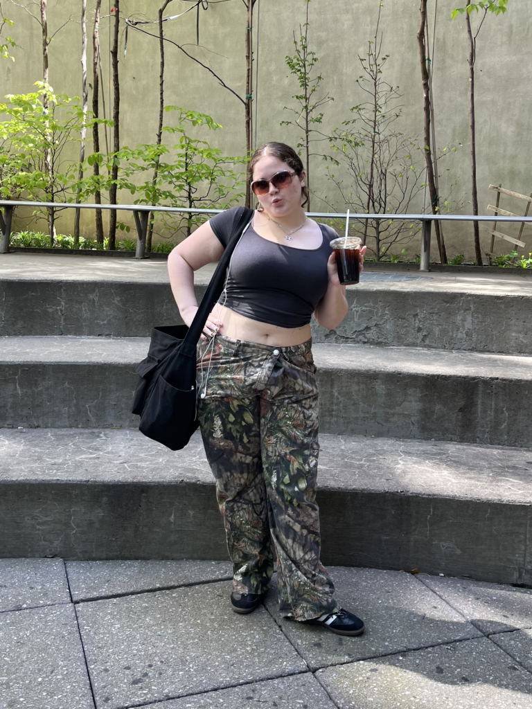 Student stands in Lang courtyard holding an iced coffee and wearing Realtree camouflage baggy pants, Adidas Samba sneakers, a cropped black t-shirt, a black tote bag, and oversized sunglasses.