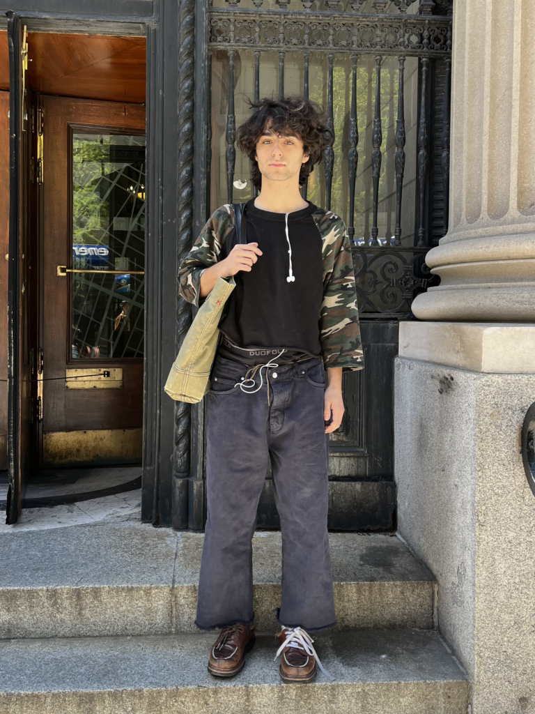 Student stands in front of a school building on Fifth Avenue wearing a raglan top with camouflage sleeves, black jeans, brown shoes, and a green tote bag