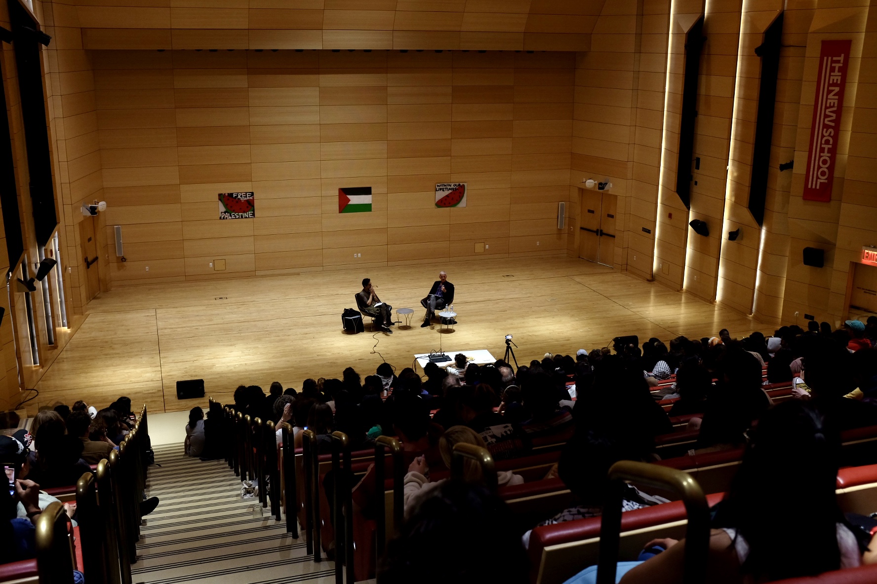 A photo of a large auditorium, taken from the back of the audience. At the center of the image is a flat stage where two individuals are seated on chairs in front of a Palestine flag.
