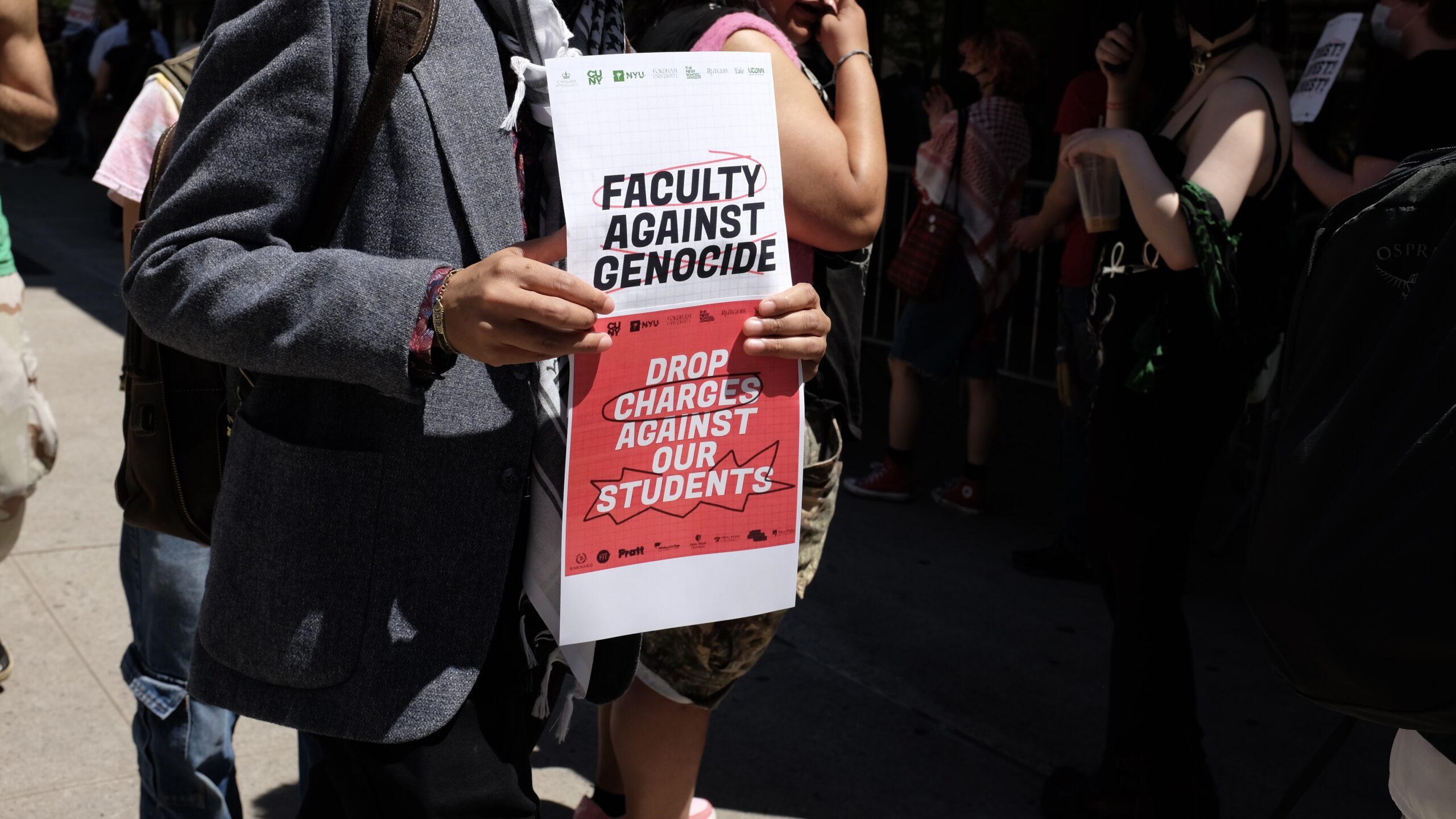 A group of people gather at a rally. One person hold up a sign that reads, “Faculty against genocide” at the top and “drop charges against our students” at the bottom..