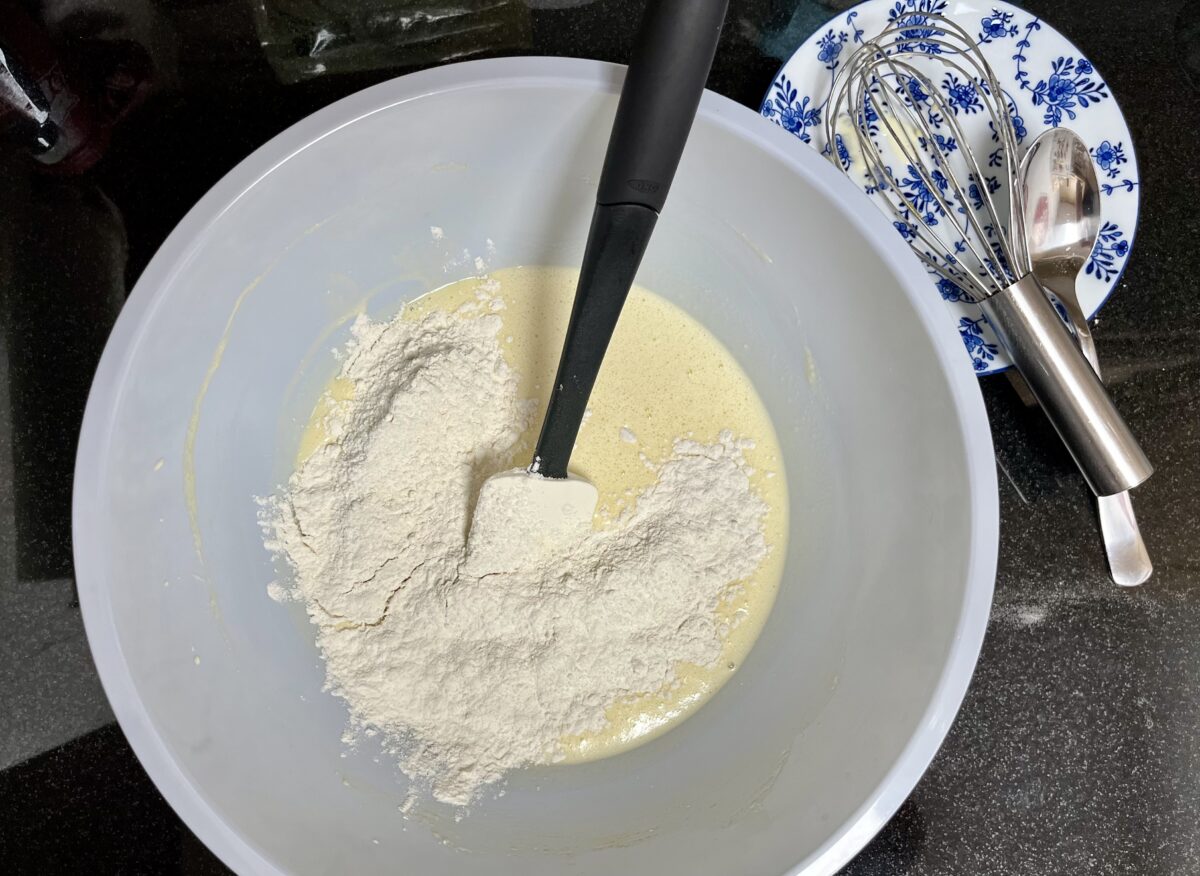 A light blue bowl filled with a pale yellow batter, flour, and a spatula sits next to a blue and white flowered plate with a whisk and spoon on top of a black countertop.