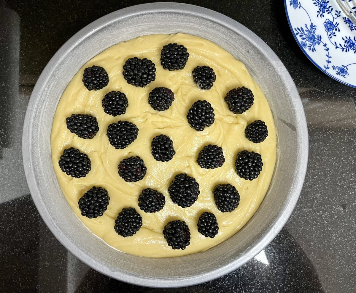 A round metal baking pan filled with yellow batter and blackberries sits on a black countertop.