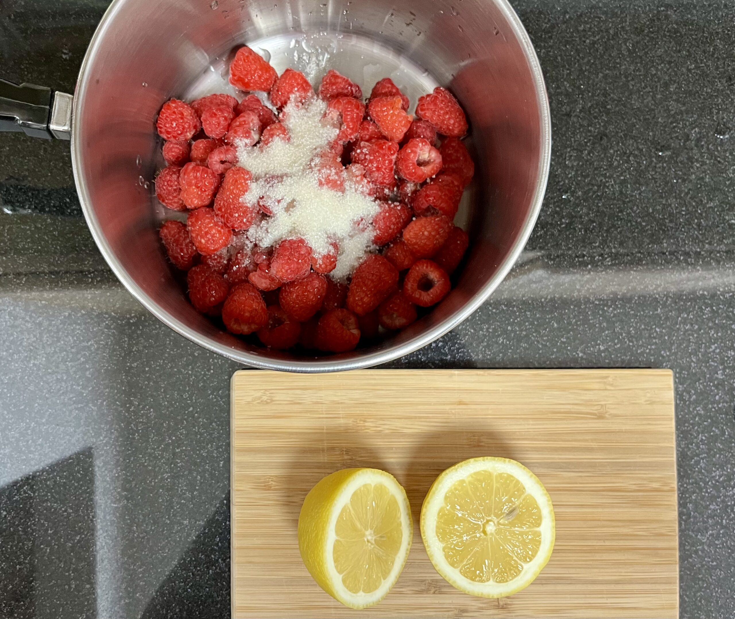 A metal pot with raspberries and sugar and a brown cutting board with a lemon cut in half sit on a black countertop.