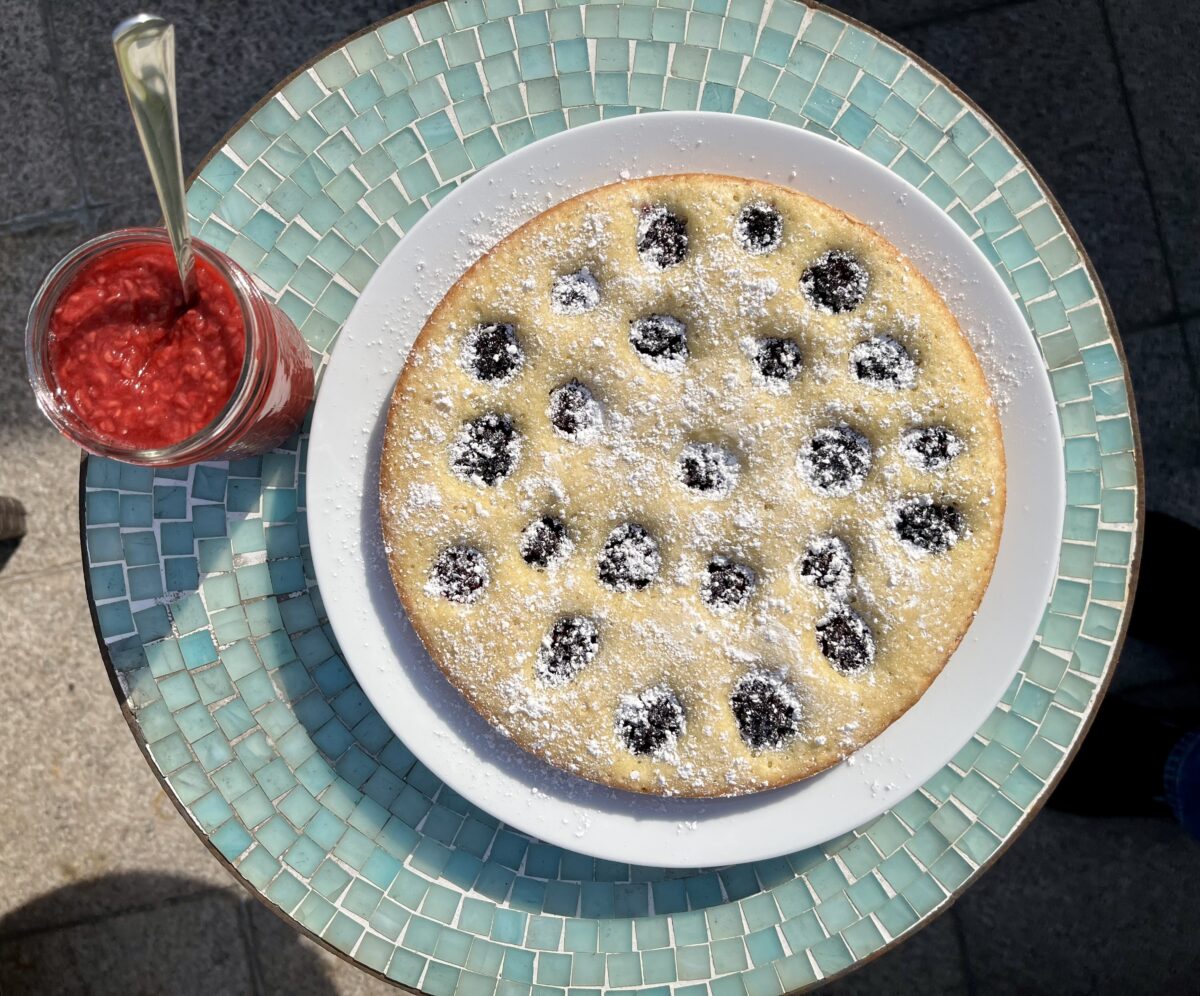 A glass jar filled with red jam and a white plate with a cake topped with blackberries and powdered sugar sit on a blue table.