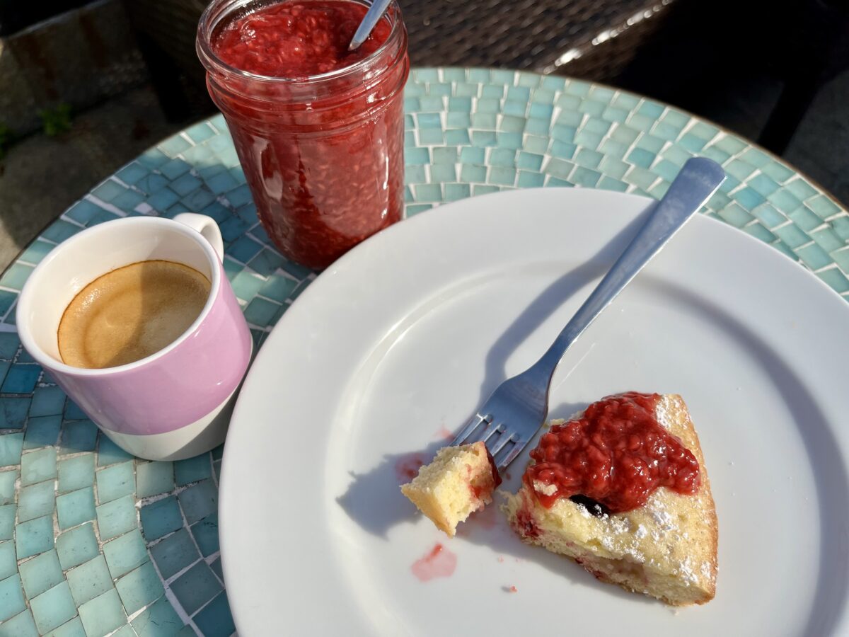 A white plate with a fork and a slice of cake topped with red jam, a purple mug of coffee, and a glass jar filled with red jam sit on a blue table.