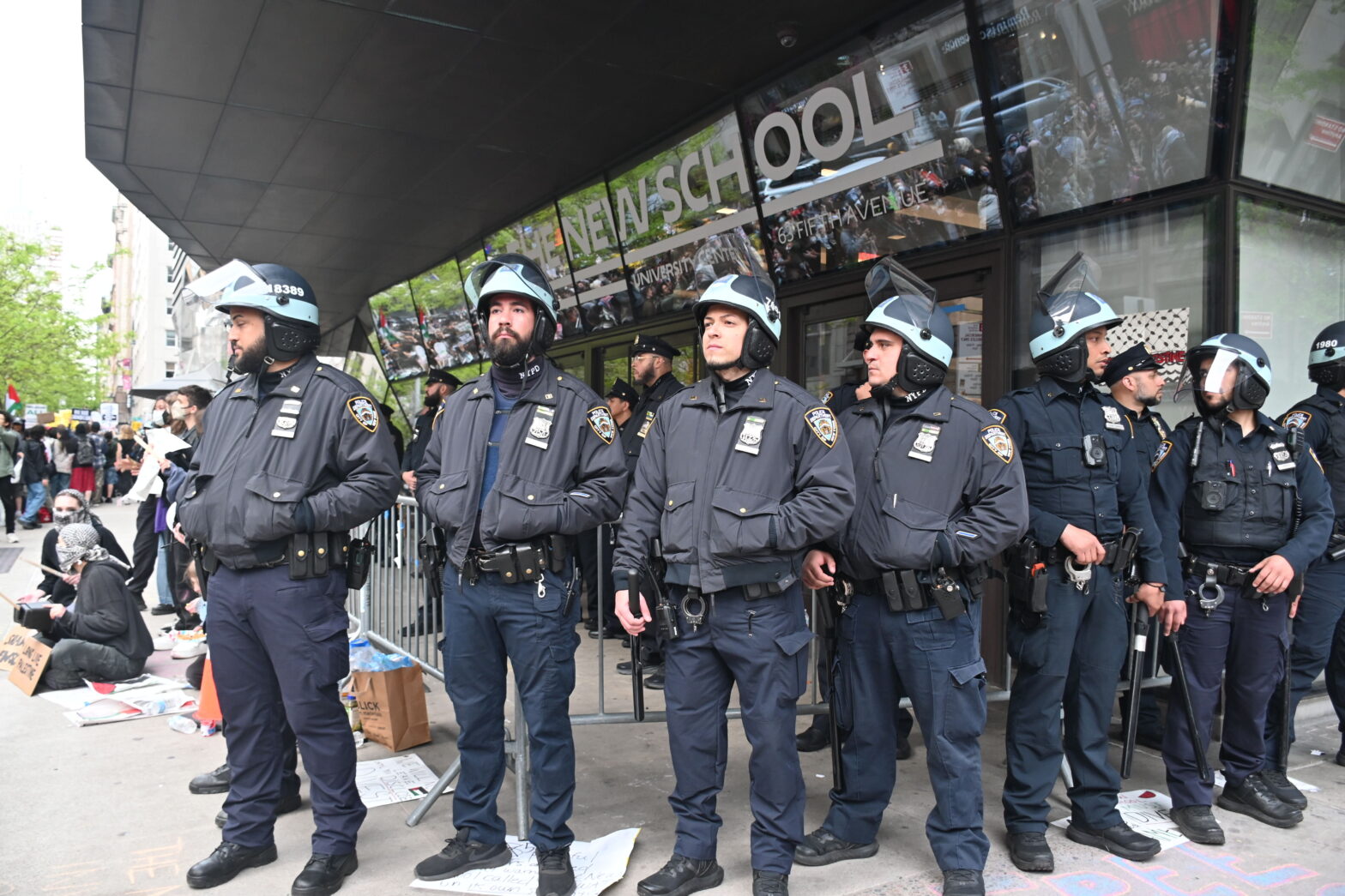 8 NYPD officers in riot gear standing outside barricades in front of the University Center. Few more officers are visible in the background, and on the right of the image, demonstrators are visible with signs.