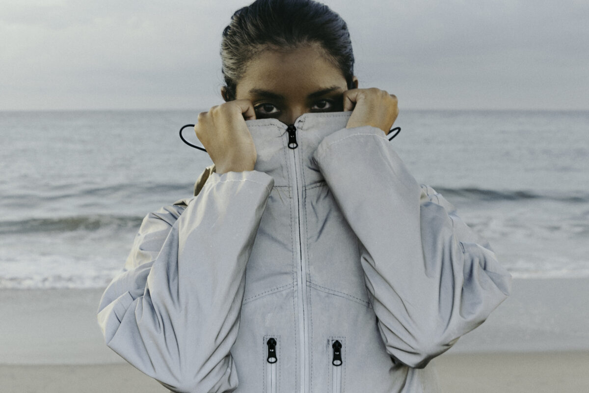 Person standing on a beach, partially covering their face with the collar of a light jacket.