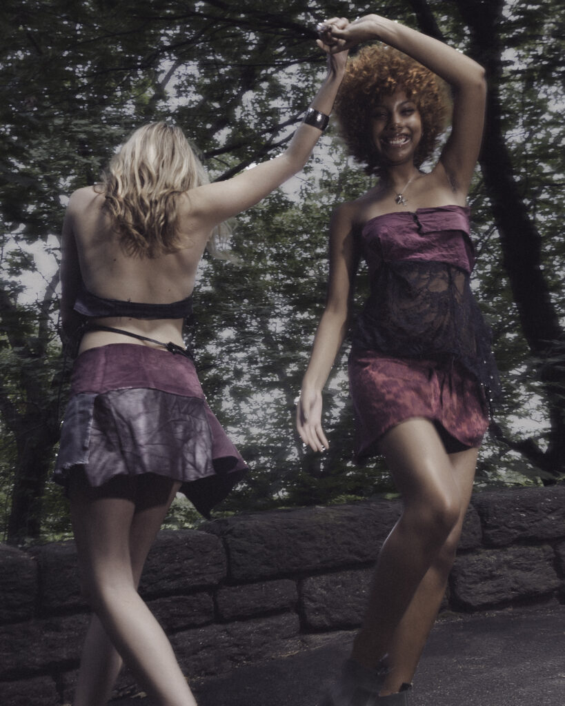 Two models smiling and dancing outdoors, wearing matching purple dresses with lace details.