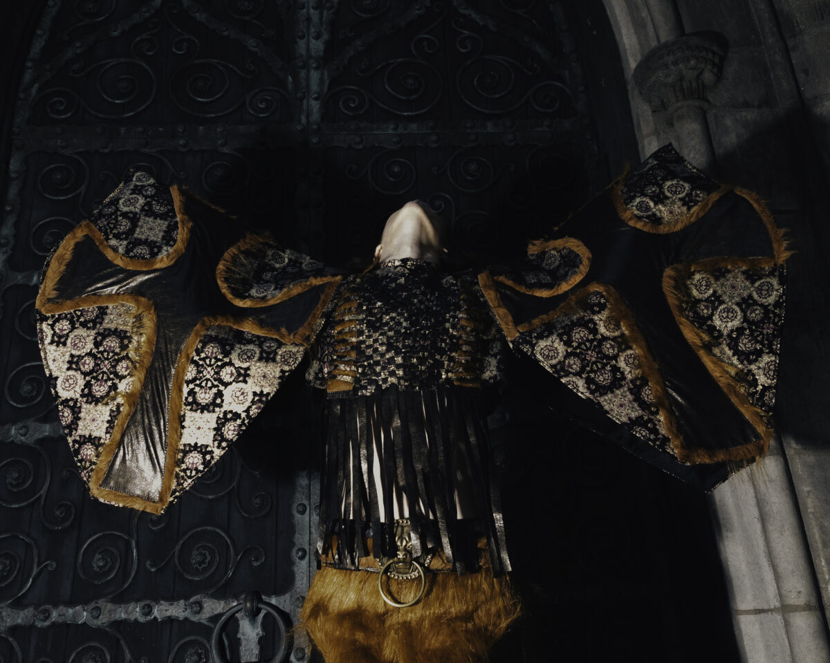 A model with arms outstretched, wearing a wing-like garment with floral tapestry, leather, and fur trim, set against a dark ornate backdrop.