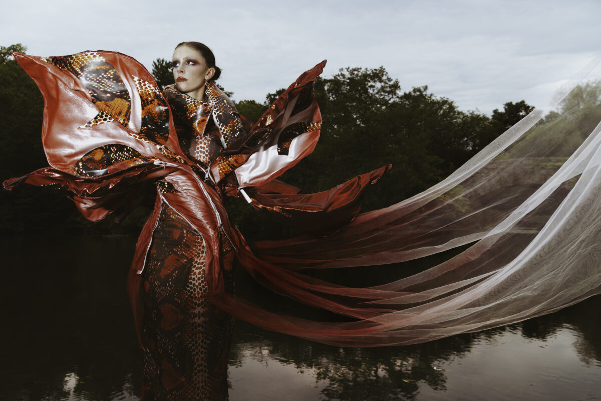 A model in a dramatic, red snakeskin-patterned gown with flowing fabrics, standing near water with trees in the background on an overcast day.