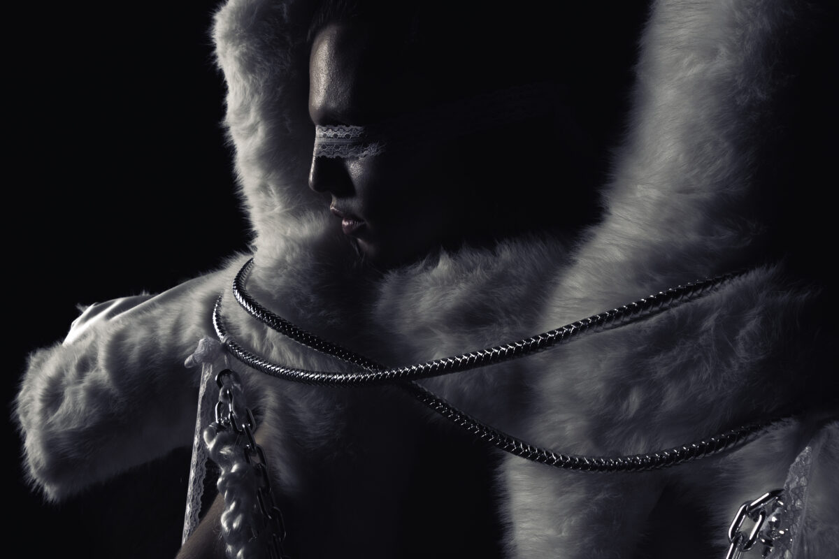 Model in a white fur garment with metallic chains wears a lace blindfold, shot in low light against a dark background.