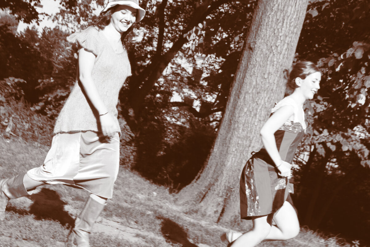 Two women running outdoors, smiling, with one in a knit top and the other in a corset-style dress, set against a backdrop of trees.