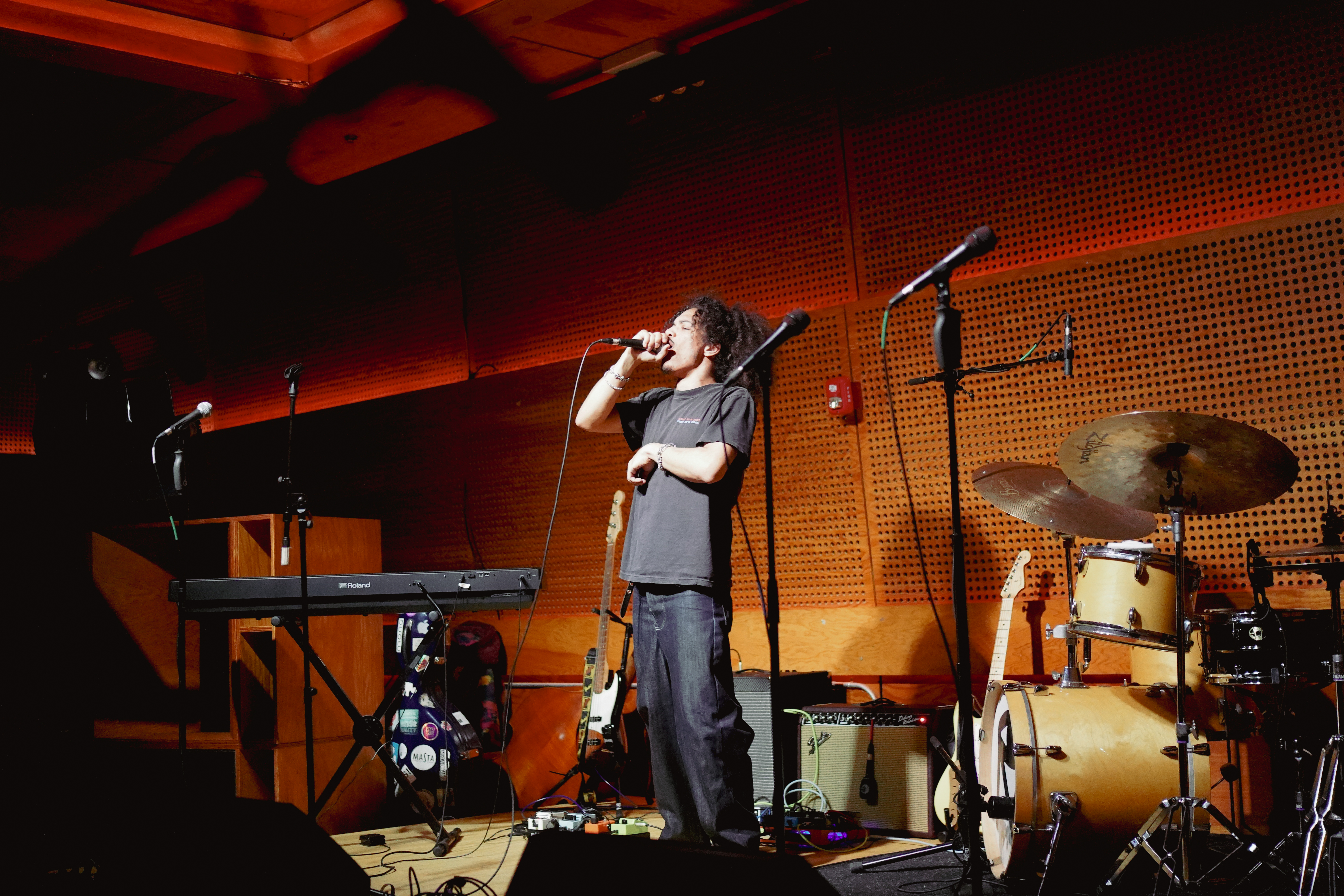 A singer in a black T-shirt performs on stage, holding a microphone with a keyboard, drum set, and amplifiers around him.