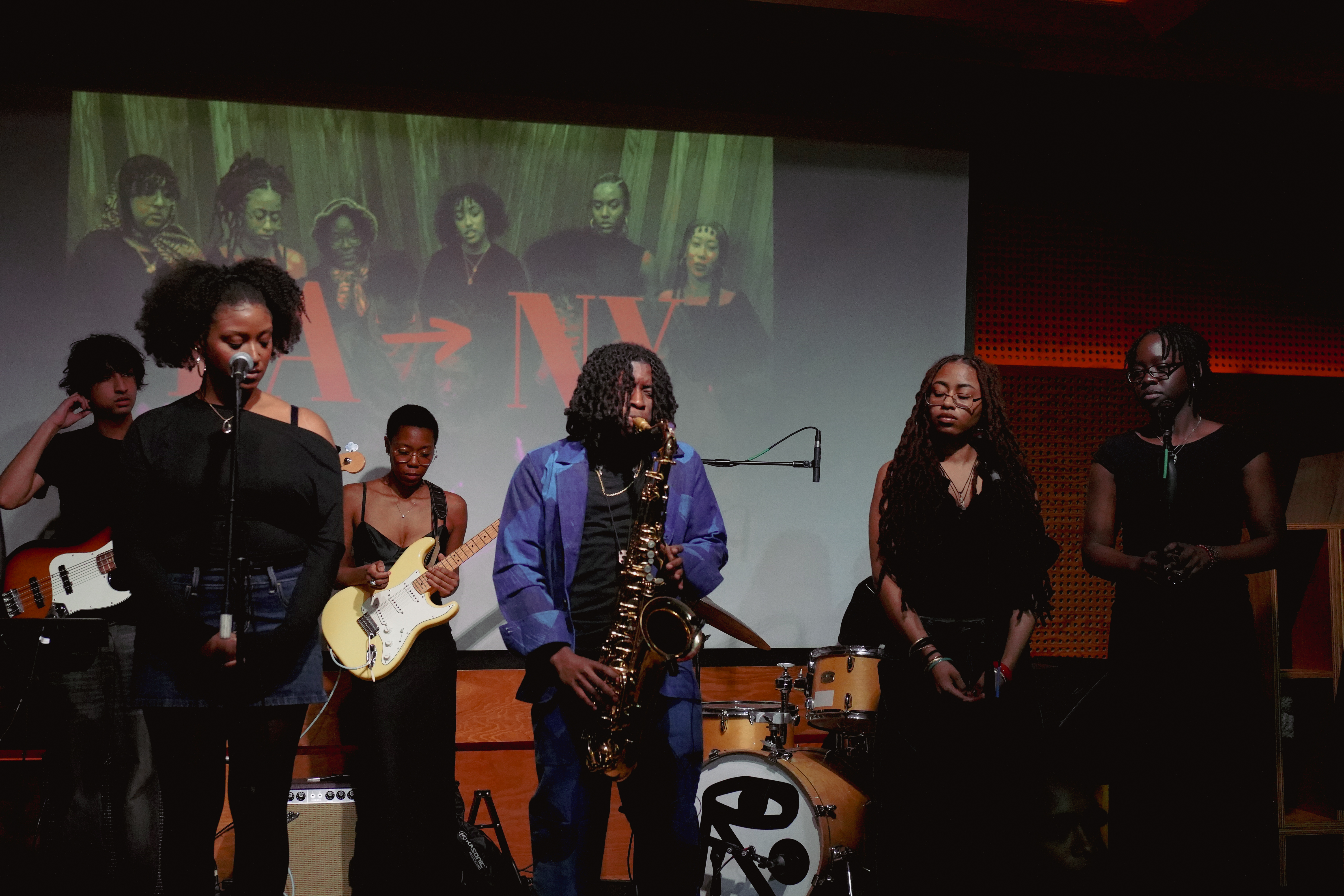 A group of musicians perform on stage, featuring a saxophonist, singers, and instrumentalists, with a projected image reading "LA - NY" in the background.