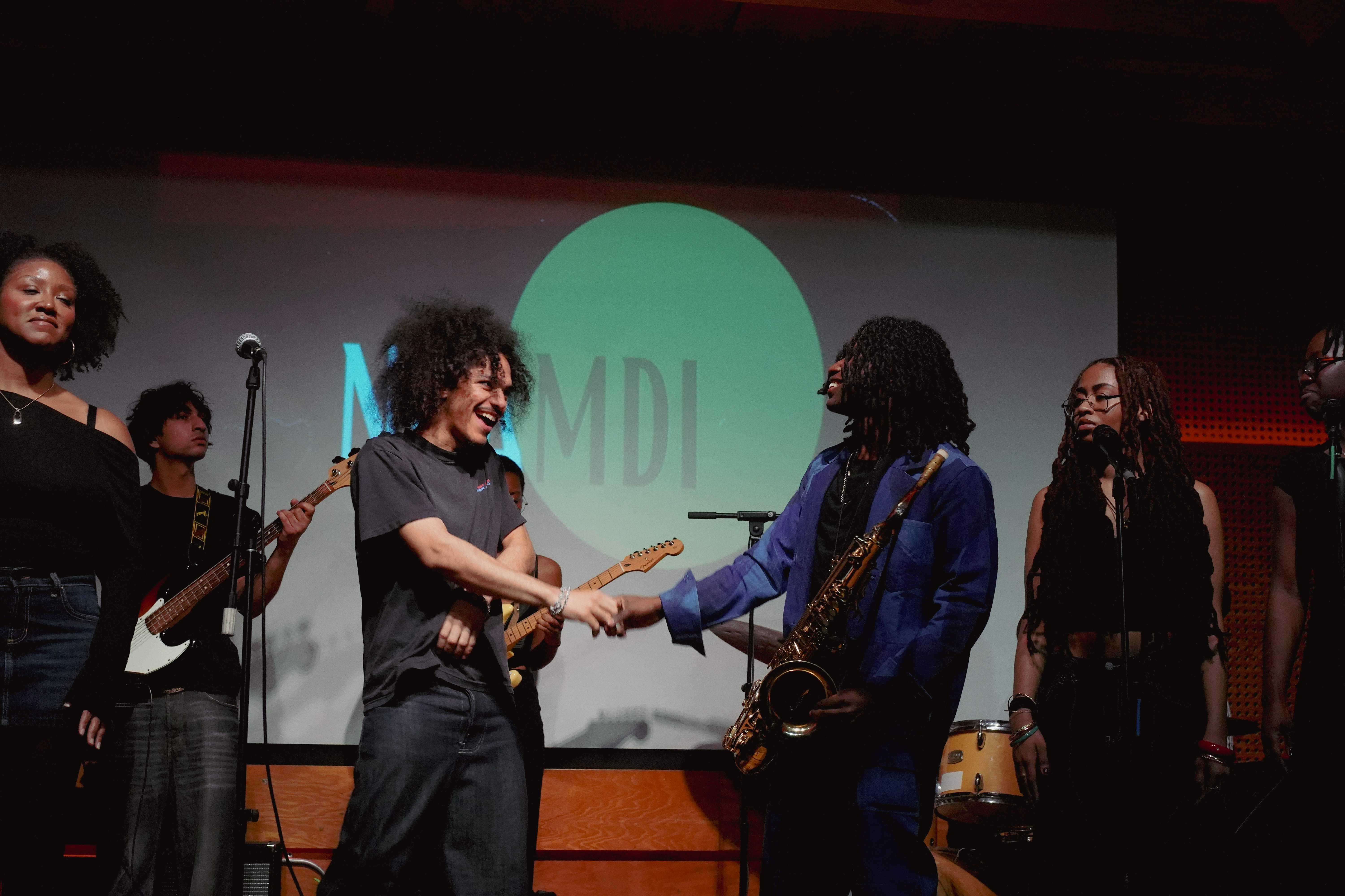 Two musicians shake hands on stage, one holding a saxophone, with a band performing in the background