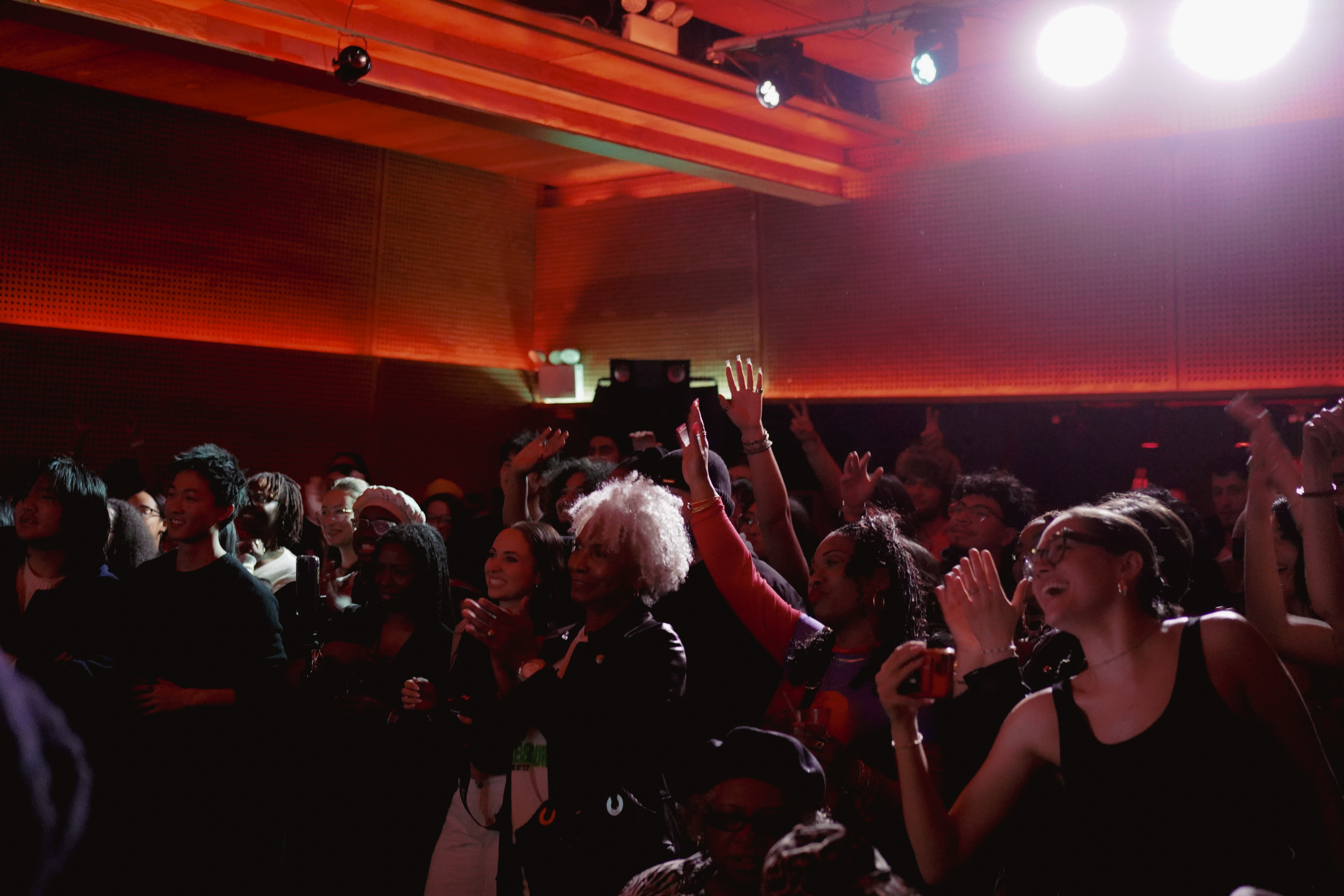 A lively audience at a concert raises their hands, claps, and takes photos under warm red lighting.