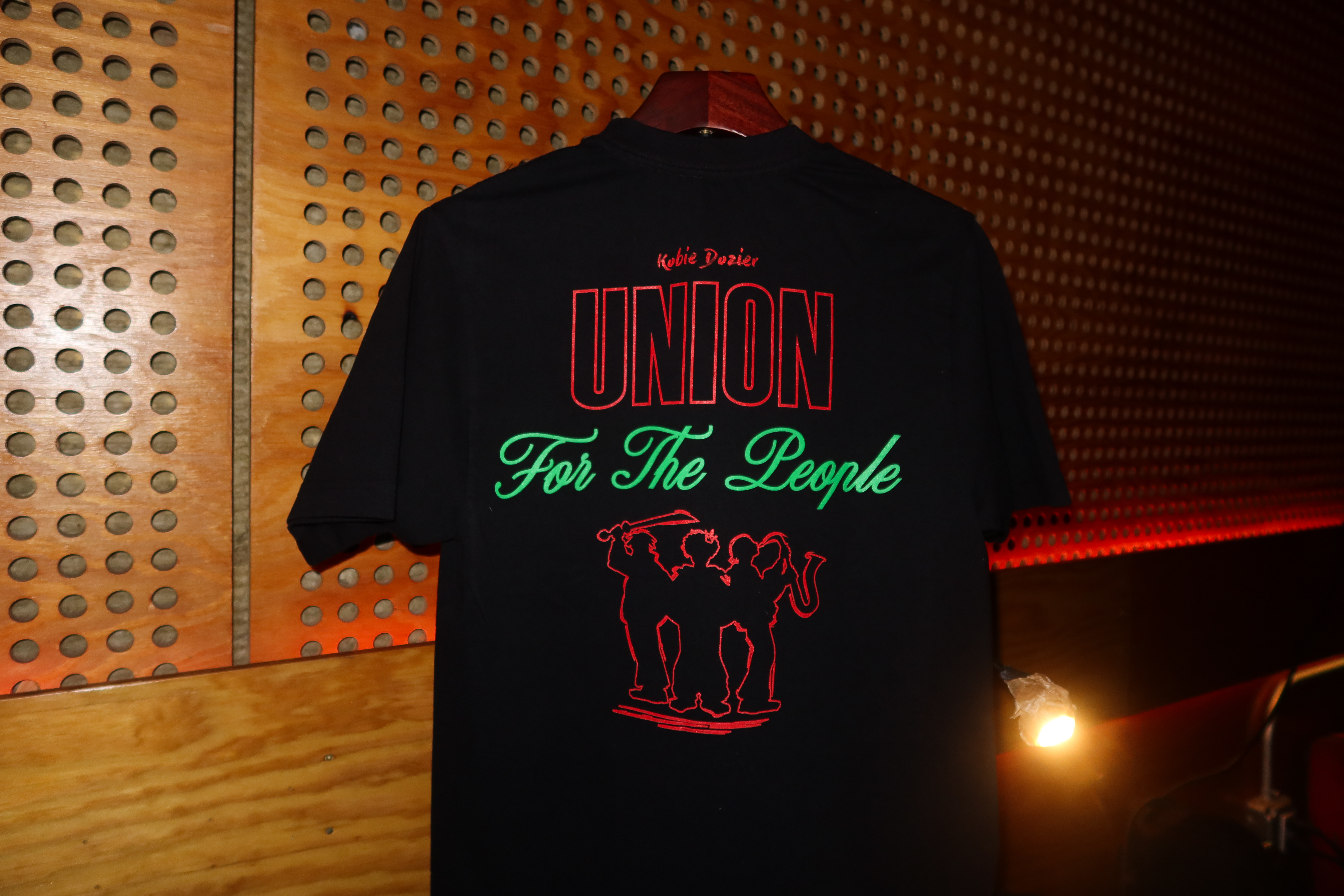 A black T-shirt hanging on a wooden wall features the text "Kobie Dozier UNION For The People" with a red outline of three musicians.