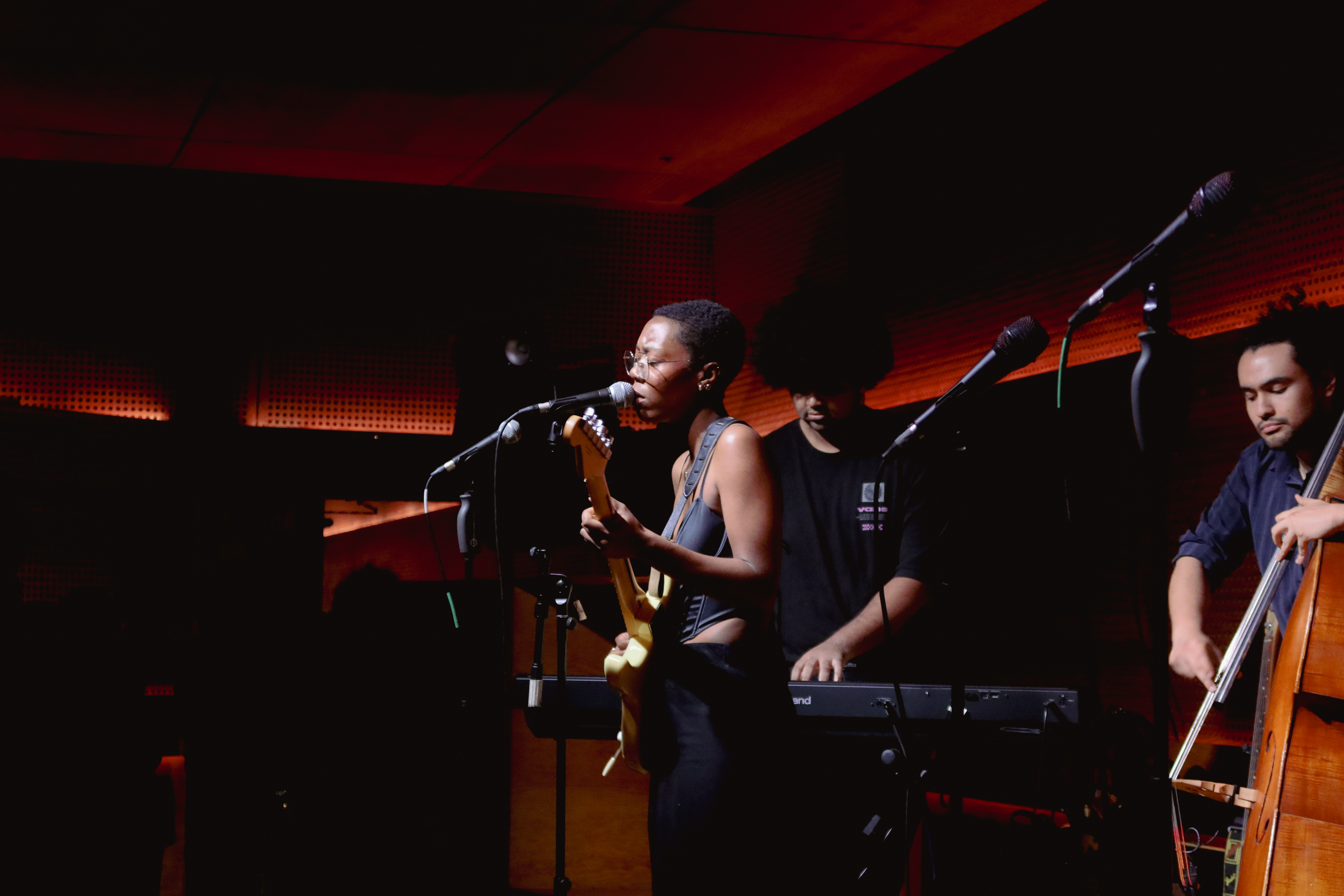 A musician sings into a microphone while playing an electric guitar, accompanied by a keyboardist and a double bass player.