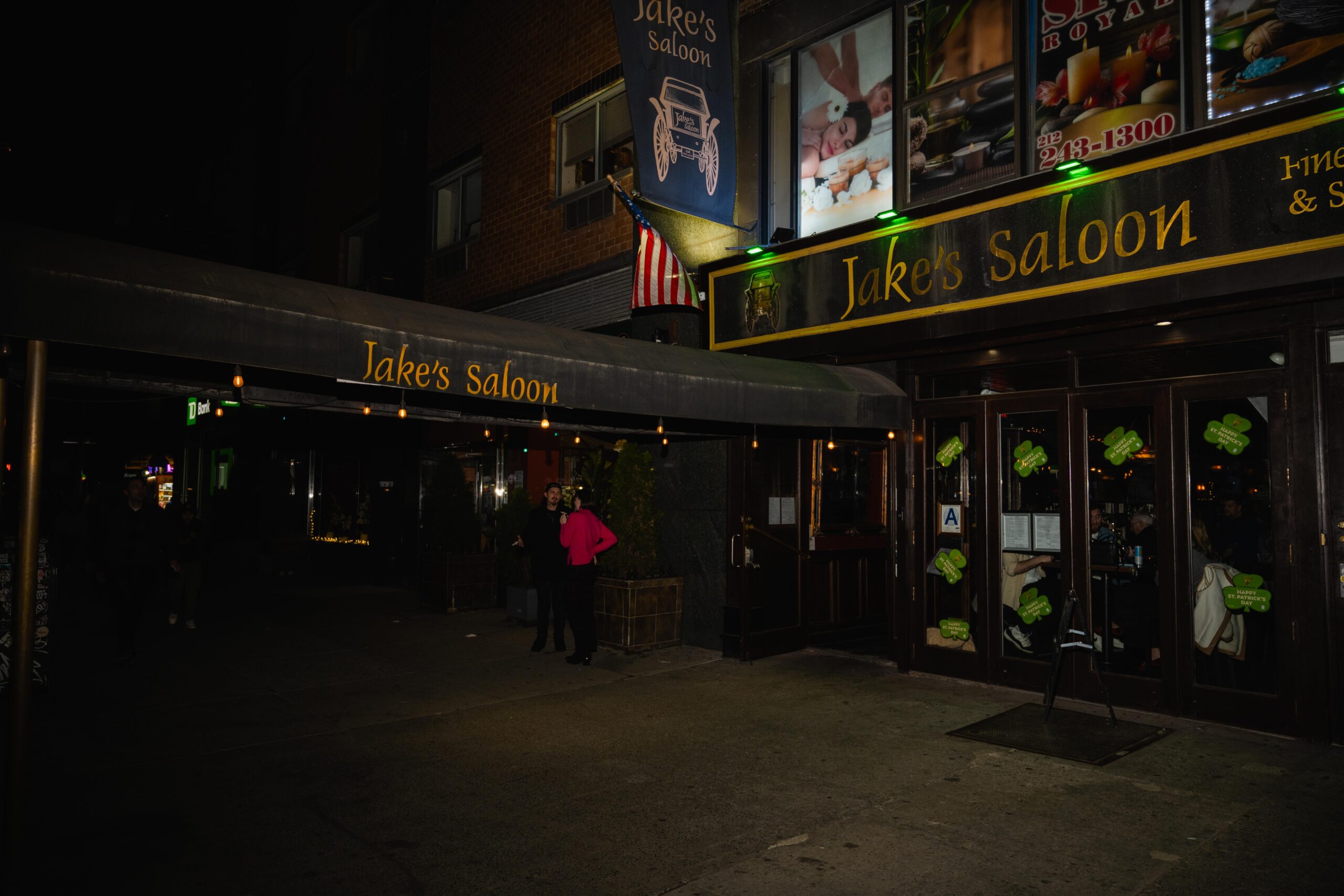 Entrance to Jake’s Saloon Chelsea. Sign and scaffolding with “Jake’s Saloon” written across. Two bar-goers standing in the front entrance.