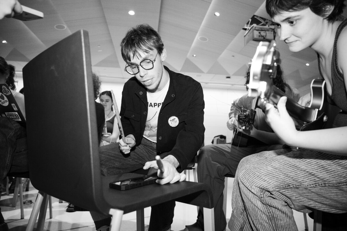 Two people look at a phone resting on a chair in the foreground. The person on the left holds drumsticks, the person on the right has a bass. The image is in black and white.