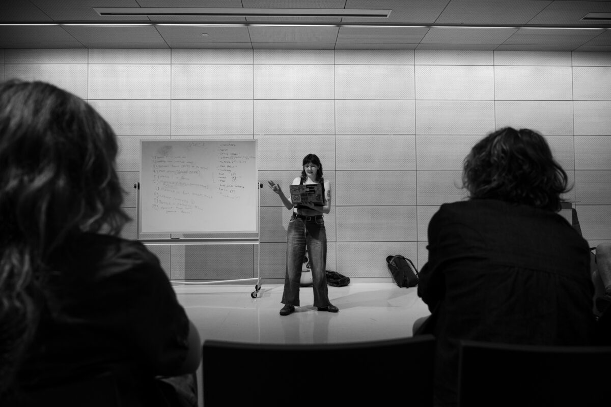 A person in jeans and a T-shirt, wearing glasses, stands in the center of the frame holding a laptop. In the background, there is a wood-paneled wall and a whiteboard with lists, and in the foreground, two heads with long hair. The image is in black and white.