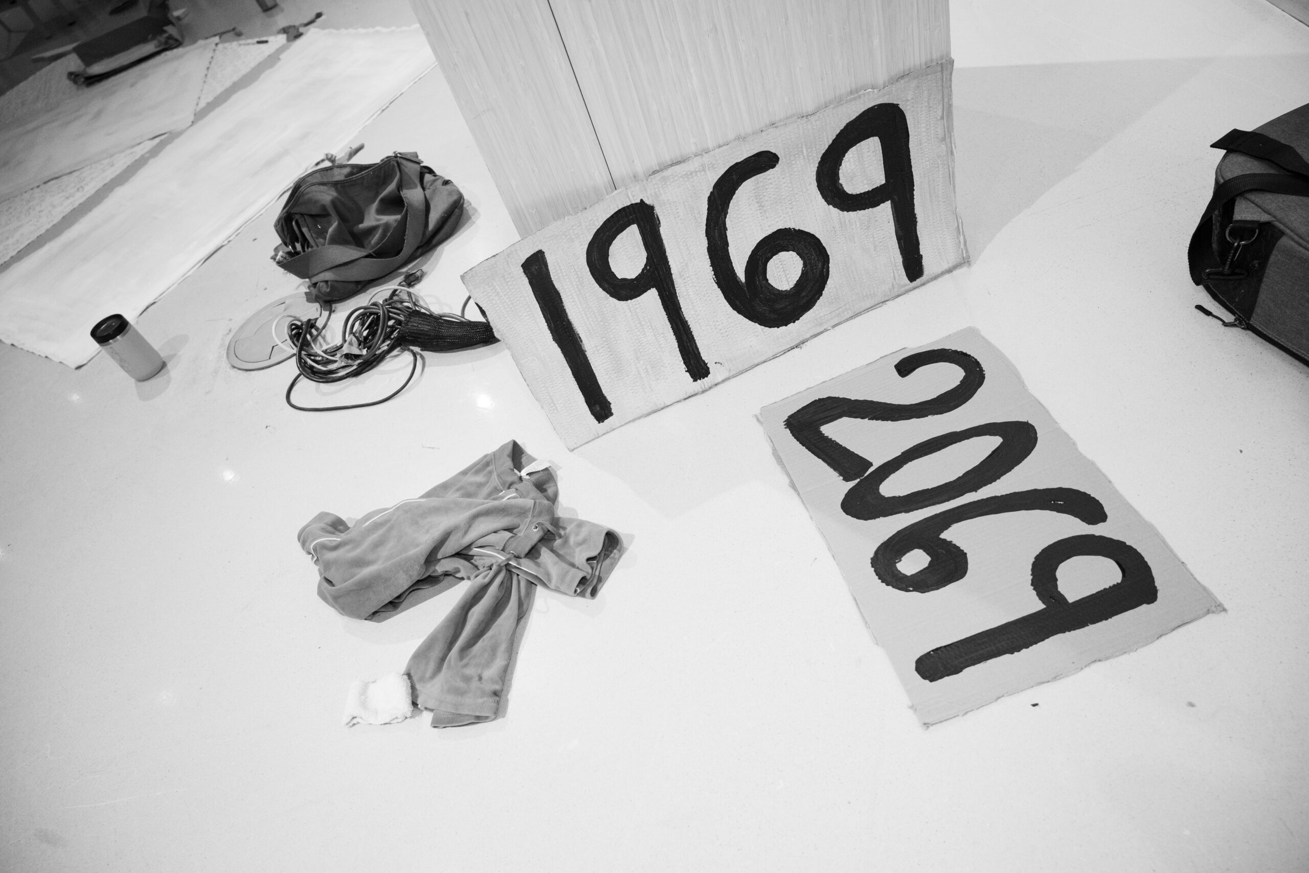 Two cardboard signs rest on the ground, next to a sweatshirt and a tangle of cables, reading “1969” and “2069” respectively. The image is in black and white.