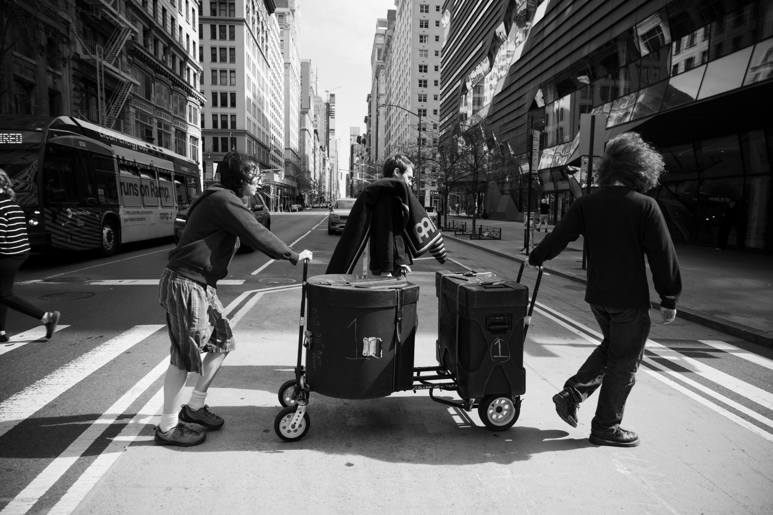 Three people push a cart holding large drum cases across a crosswalk. Large buildings are visible in the background. The image is in black and white.
