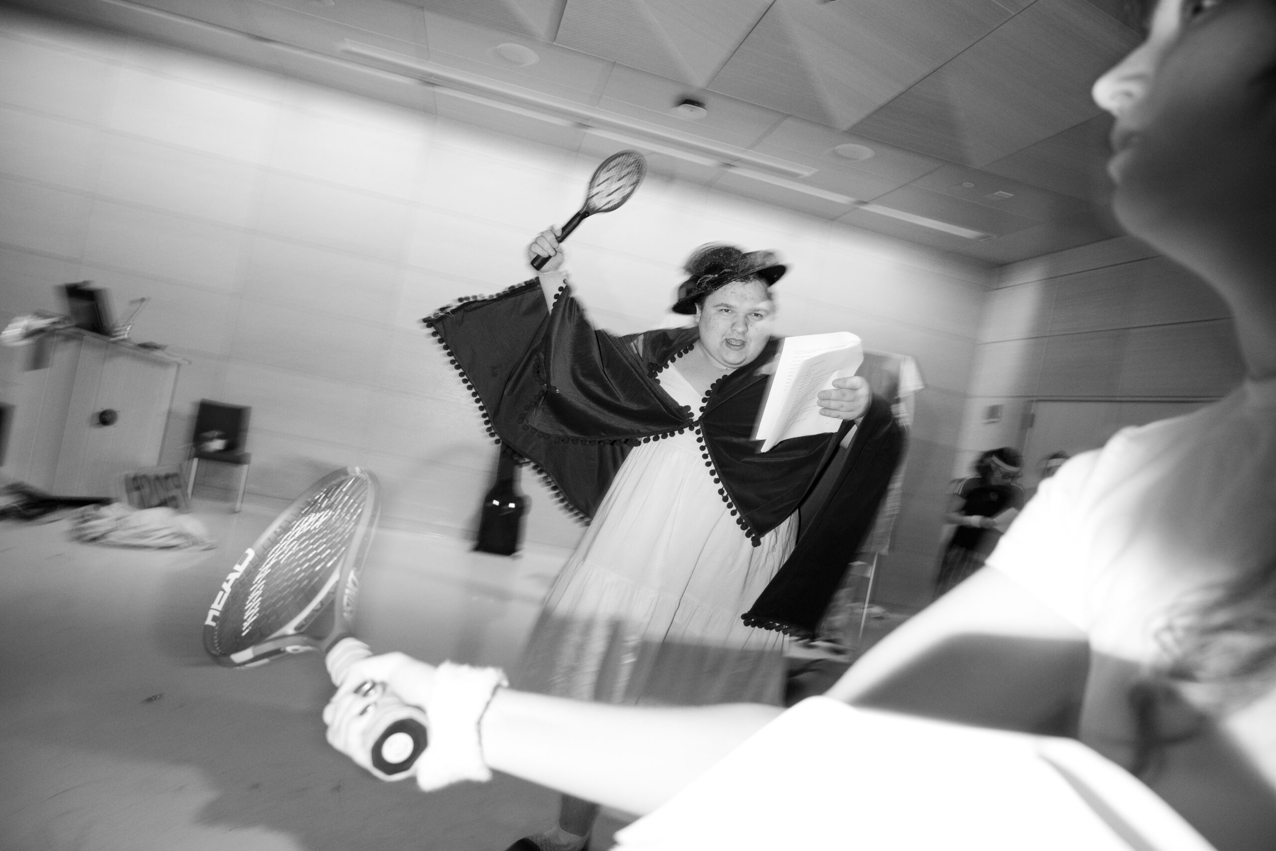 A blurred, in-motion photo of a person in a cloak, holding a script and a tennis racket in the center of the frame. In the foreground, there is the arm of another person holding a tennis racket and a script. The image is in black and white.