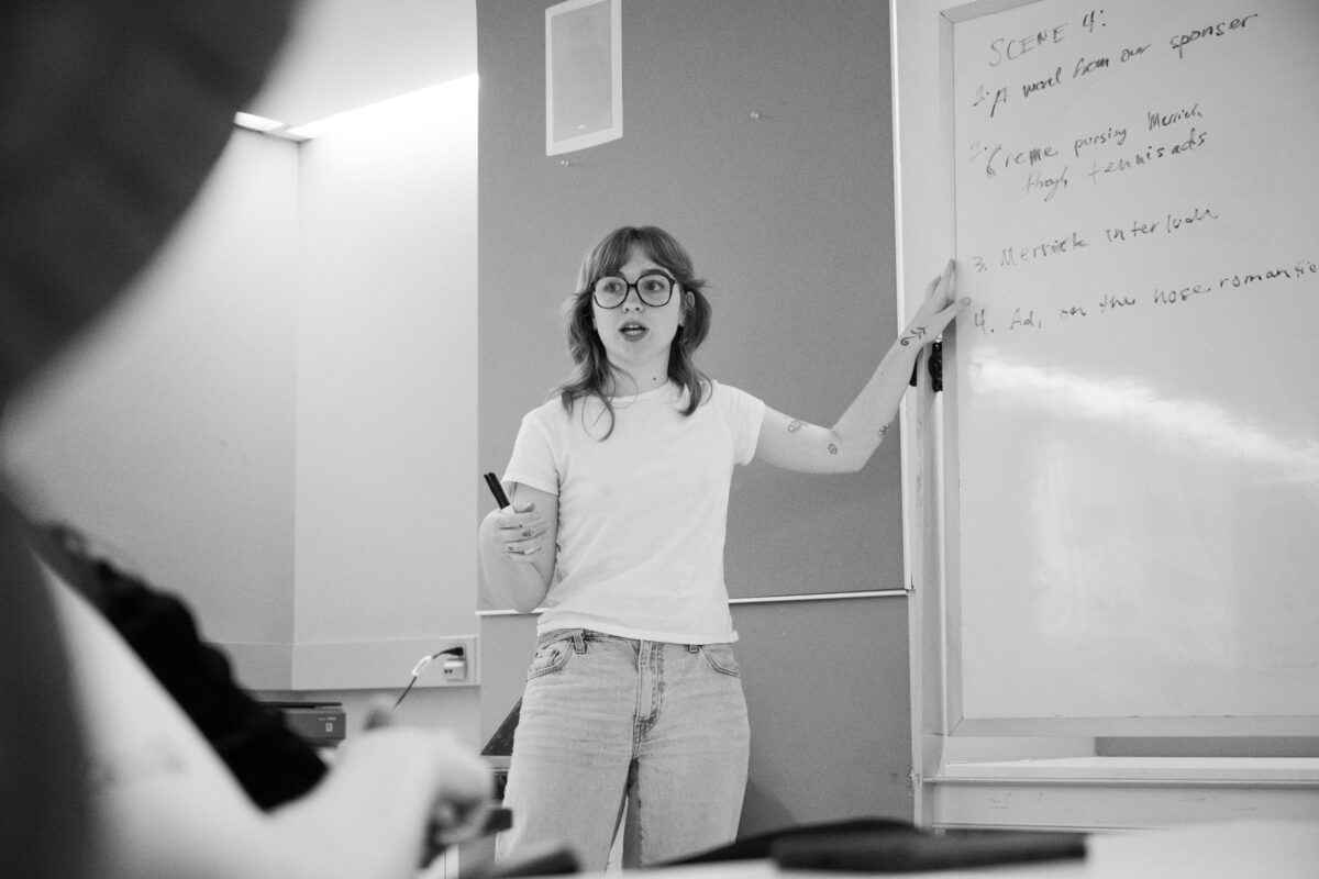 A person stands in the center of the frame in jeans and a T-shirt, gesturing at a whiteboard with a list of moments of a scene written on it. The image is in black and white.