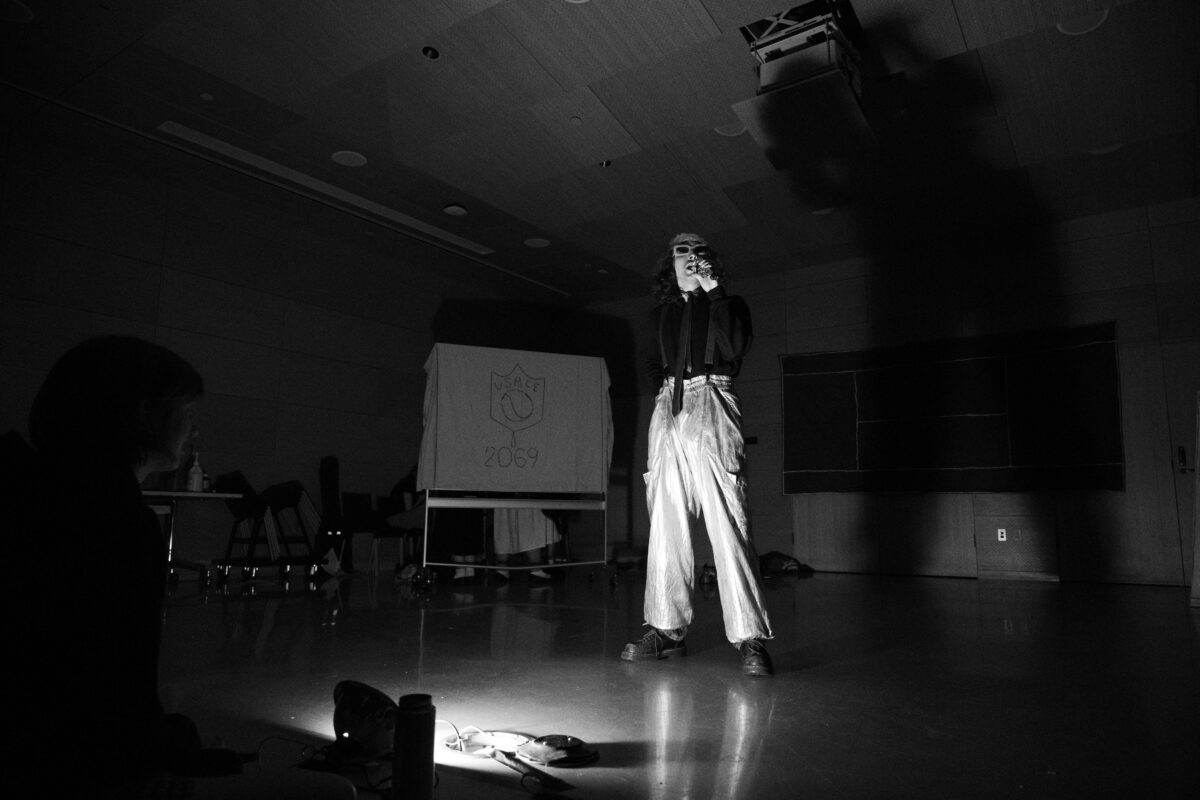 A person, wearing sunglasses and holding a fake microphone, stands in the center of the stage, lit from below. His shadow is cast on the wall behind him. In the background, a whiteboard draped in a sheet reads “U.S.A.C.E 2069.”