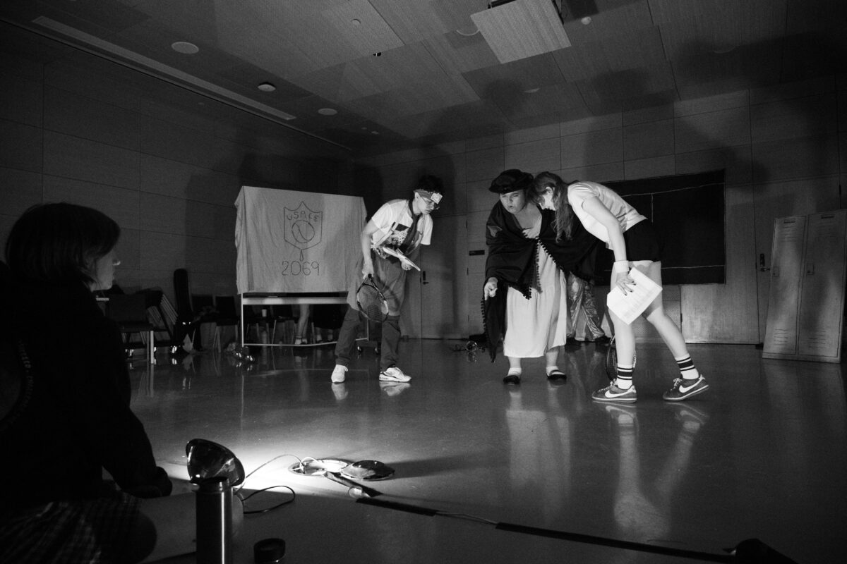 Three people stand in a circle, in the center of a large room, bent over and facing each other. They are lit from below, and their shadows are cast on the wall behind them. Behind them, a rolling whiteboard is draped in a sheet that reads “U.S.A.C.E 2069.”