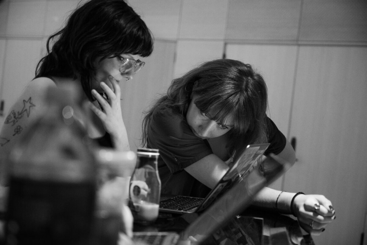 Two people look at a laptop. The image is in black and white. In the foreground, a Starbucks cold brew glass bottle.