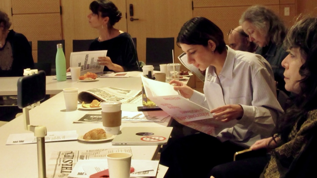 A woman in a light blue button up bends over a copy of the newspaper designed for the event. She is one of several women seated around a long white table, the top laden with croissants and coffee cups.